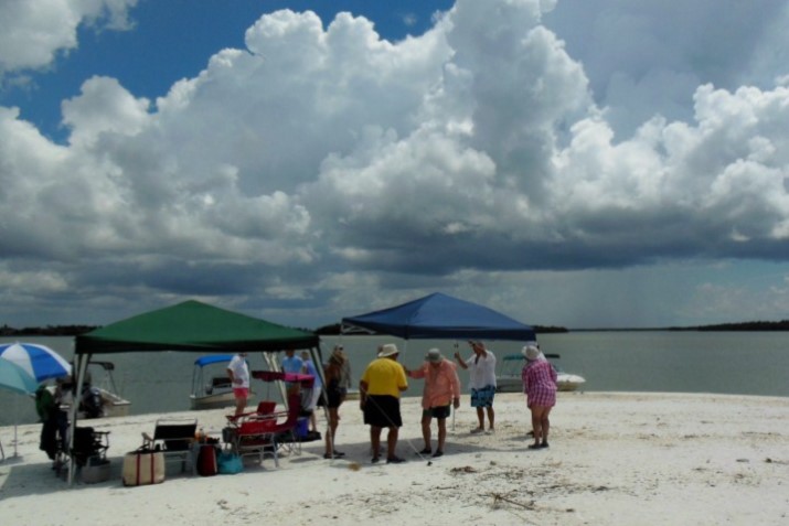 Photo, beach picnic with several adults under and around some beach awnings and umbrellas. In background are 3 boats pulled up to the beach.